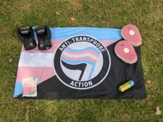 Anarchist trans flag laid out on grass; on top of it are boxing gloves, pads, an energy drink and a packet of face masks.